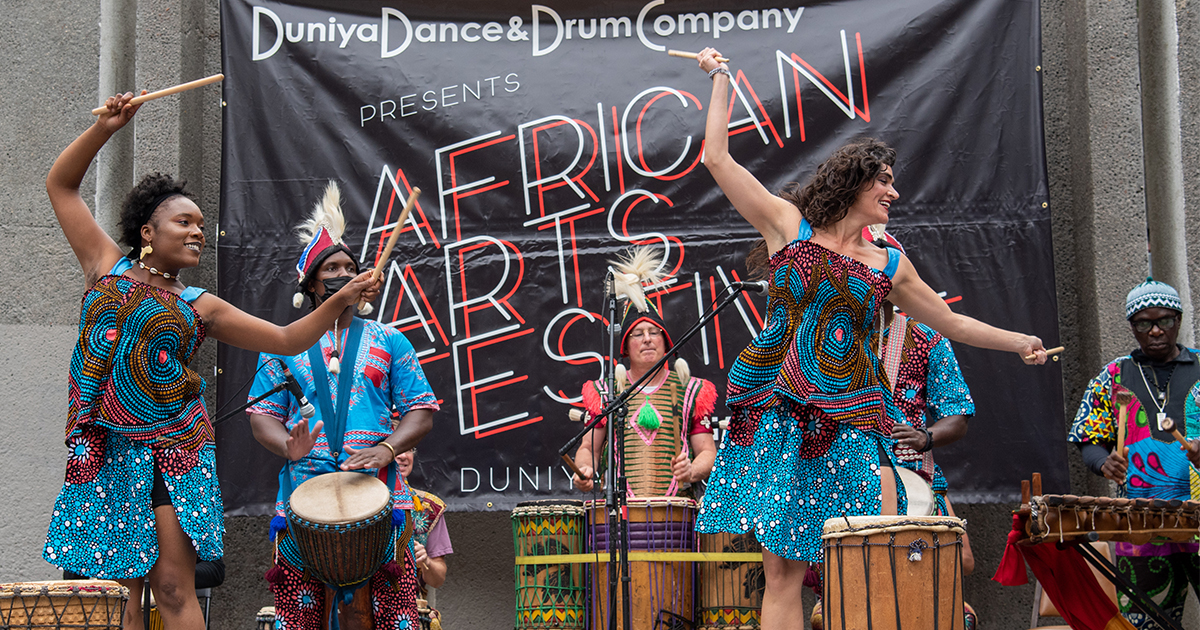Dancers and drummers in colorful African regalia, smiling and performing on an outdoor stage in front of an "African Arts Festival" banner