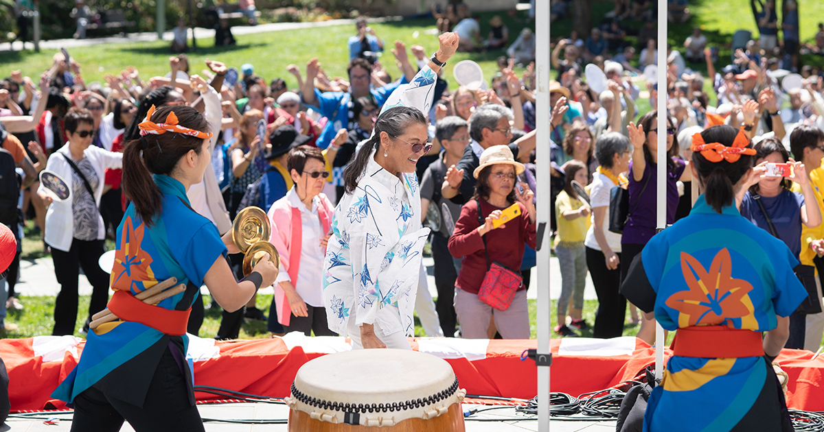 Taiko drummers and bon dancers performing and leading the crowd of people behind them outdoors on a lawn