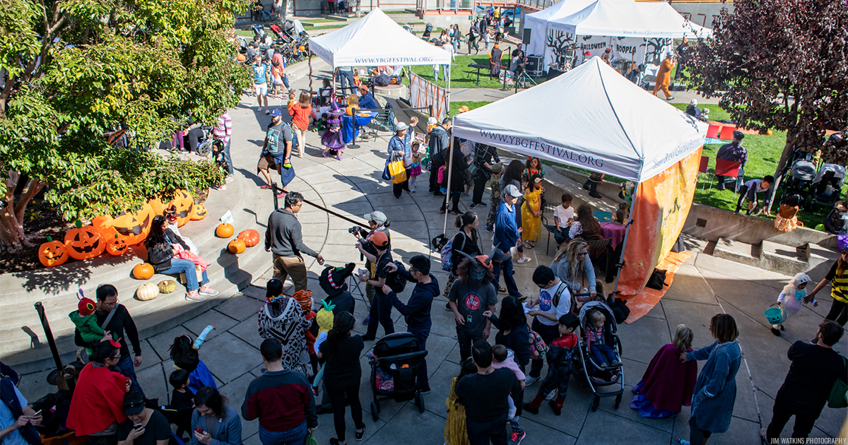 Top view of a large crowd of kids and families dressed in costumes walking outside across the Children's Garden with tents and pumpkins and other Halloween decoration set up