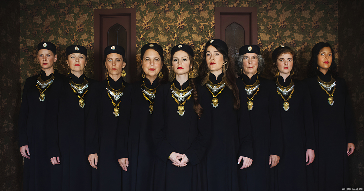 9 members of Kitka, front-facing, wearing black outfits and gold necklaces, standing in front of a green and gold patterned wall