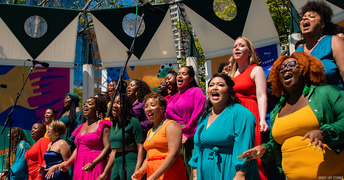 Members of CO-LLAB Choir each dressed in different, vibrant solid colors, singing passionately on an outdoors stage.