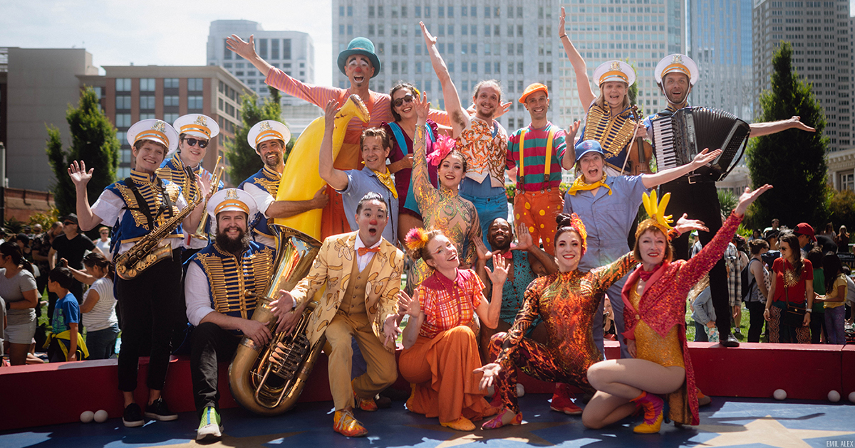 Members of Circus Bella wearing bright pink, yellow and white circus costumes, smiling and front-facing outdoors, audiences and a cityscape behind them.