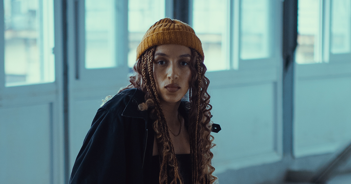 Jada Imani wearing a yellow beanie and black clothing, front-facing, in front of a white wall and row of windows.