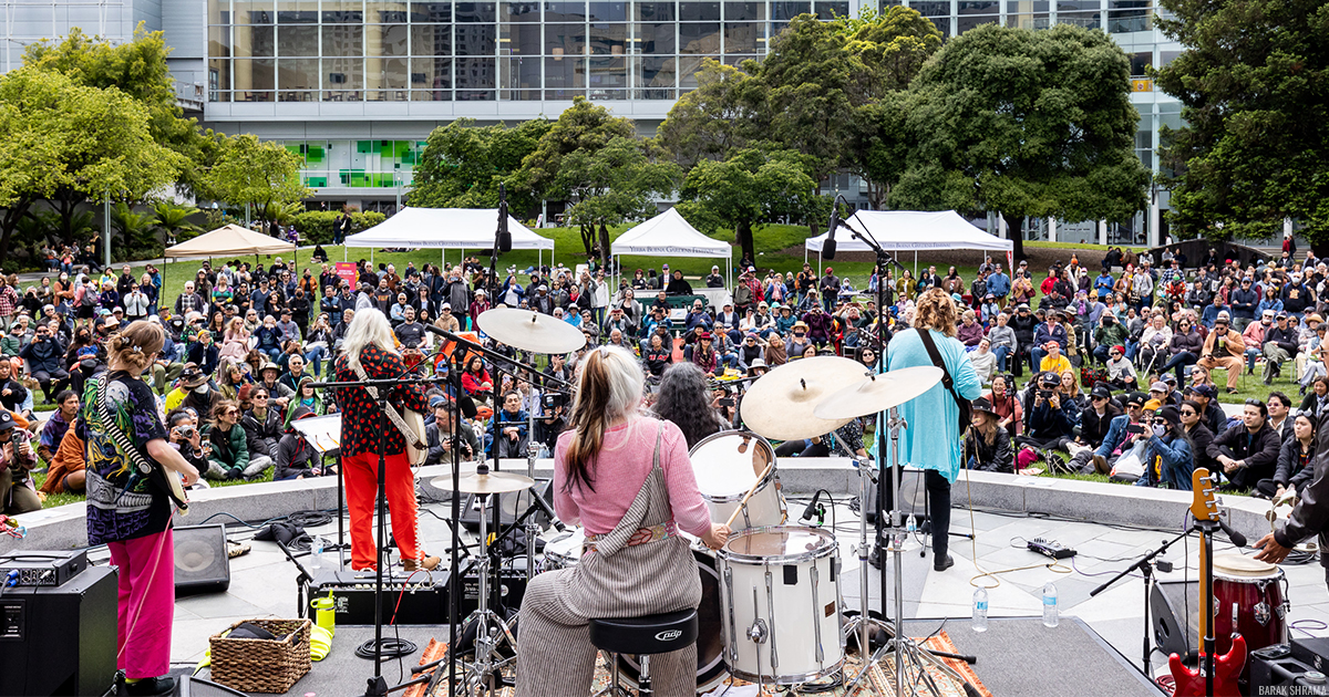 A photo from behind a music band playing on a outdoor stone stage, in front of a large audience sitting on the Yerba Buena Gardens lawn