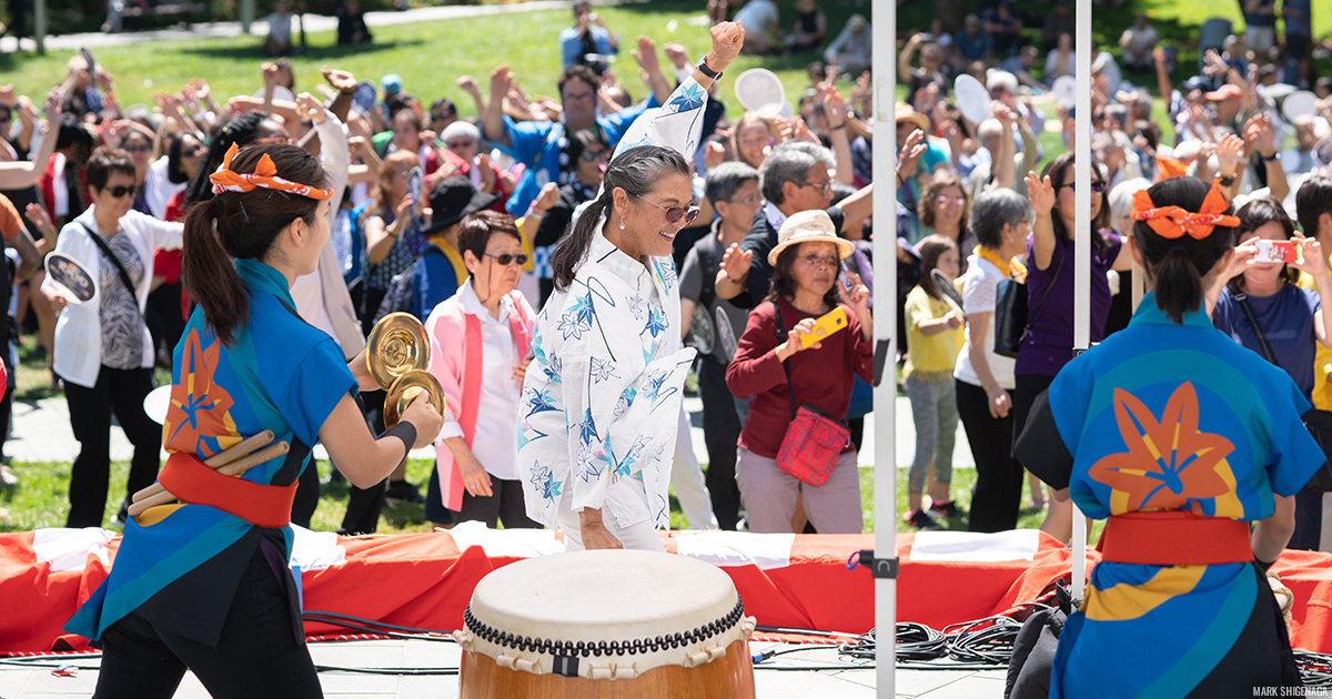 Taiko drummers and bon dancers performing and leading the crowd of people behind them outdoors on a lawn