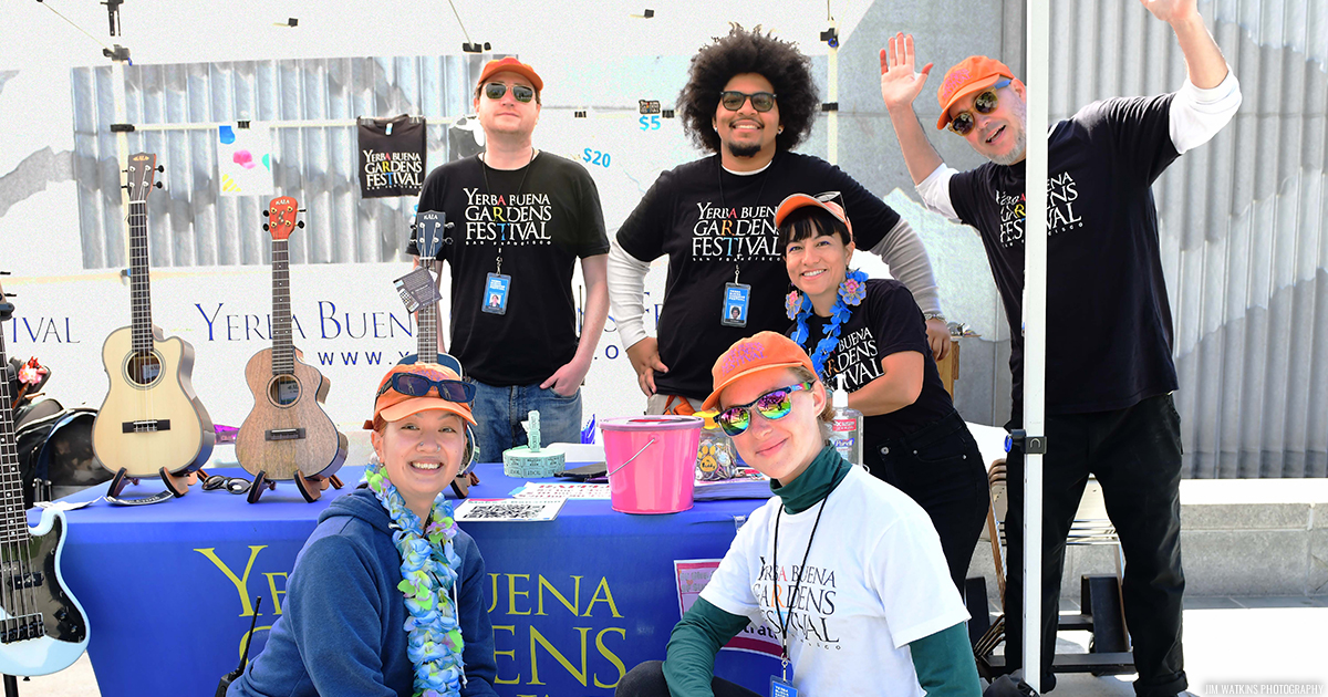 A small group of Event Staff wearing YBG Festival t-shirts and orange hats, smiling and standing gathered at an information table