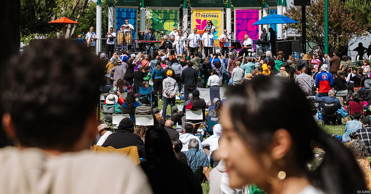 051725_JoeBataan_DSC00417-2_Credit-NixGuirre-copy Two unfocused, smiling people in the foreground, a clearly large audience in the background watching a concert on a sunny Yerba Buena Gardens Festival stage.