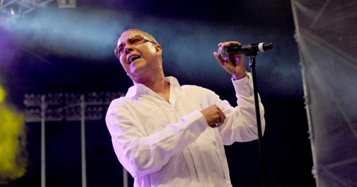 Hermán Olivera wearing glasses and a white shirt, holding a microphone on a stand in one hand as he turns out to the left.