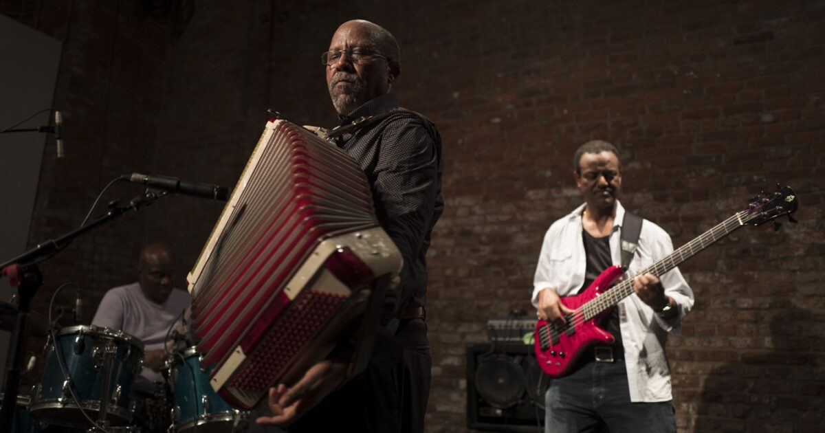 Hailu Mergia playing the accordion with a guitarist and drummer playing behind him against a brick wall.