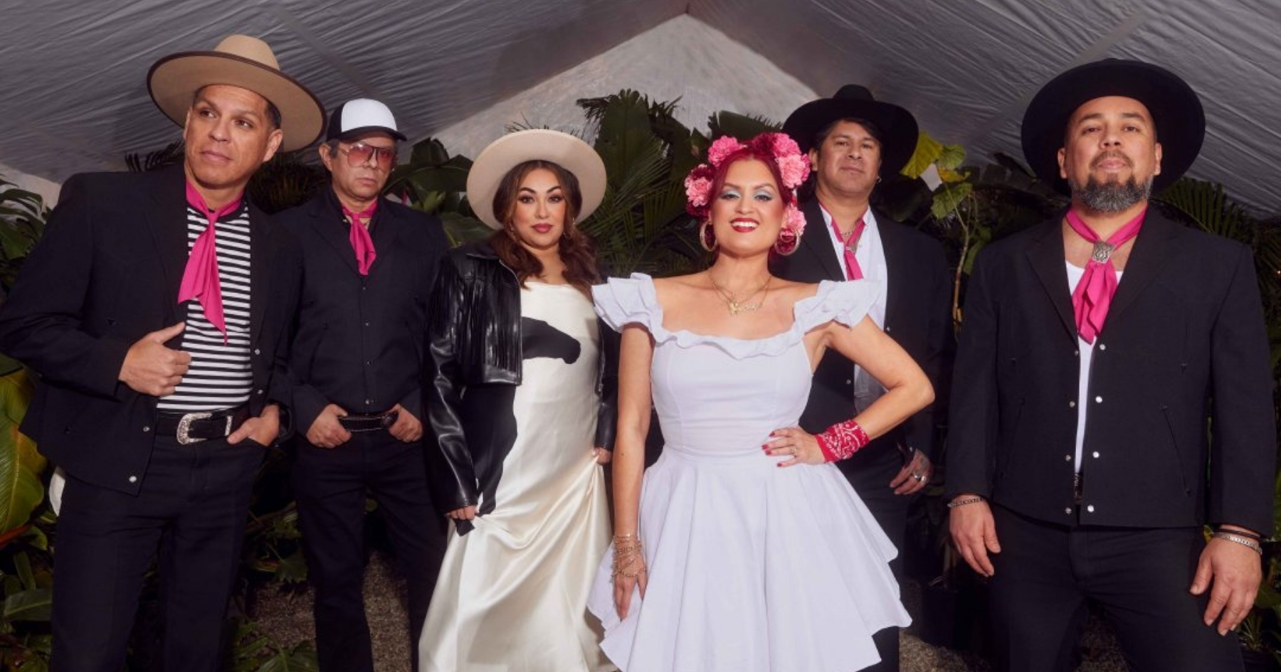 Members of Las Cafeteras, wearing black, white and pink outfits, front-facing, standing under a white tent.