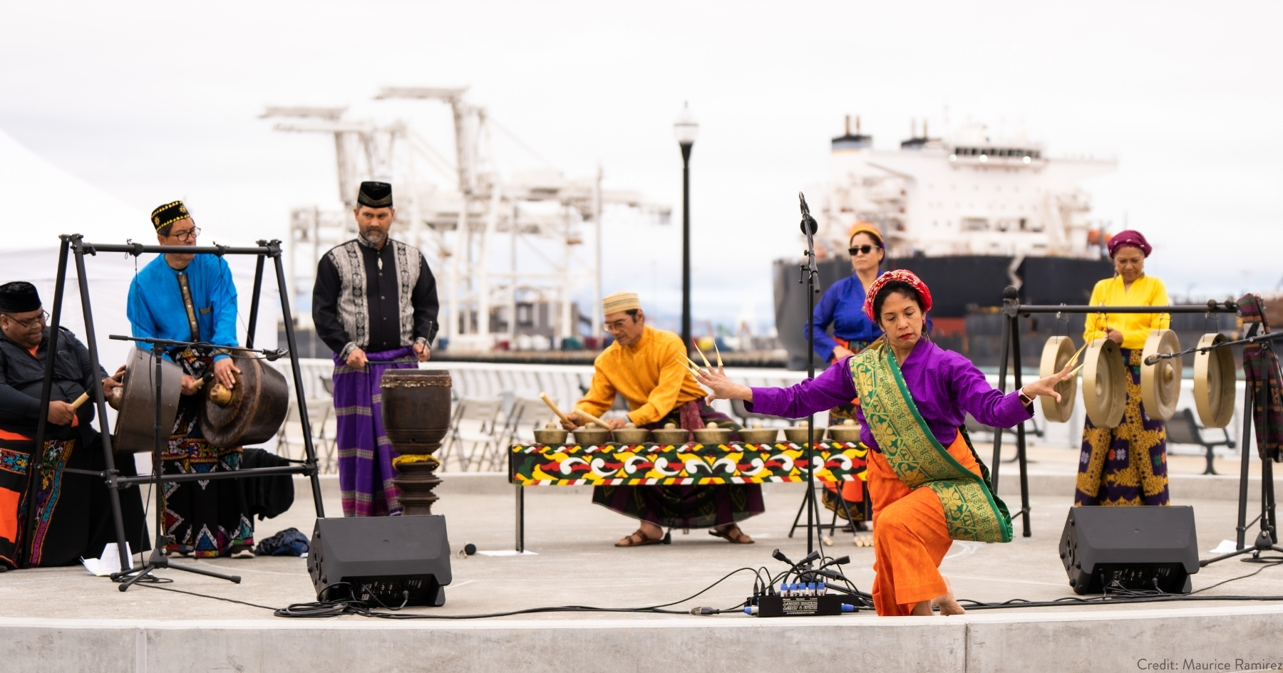 A group of 7 performers in traditional Filipino attires—6 are playing various gong instruments, 1 performing a dance at the front