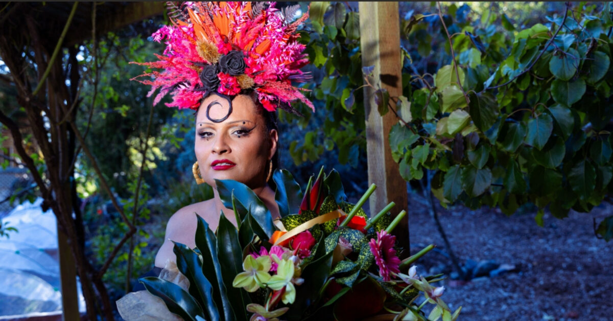 060726_PrideInYerbaBuena_1800x945_web Per Sia wearing a colorful, tropical pink flower crown, standing behind a green bouquet of plants and flowers, a dimly lit green orchard behind them.