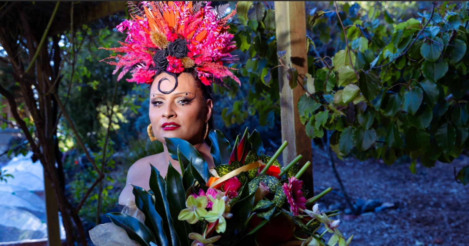 Per Sia wearing a colorful, tropical pink flower crown, standing behind a green bouquet of plants and flowers, a dimly lit green orchard behind them.