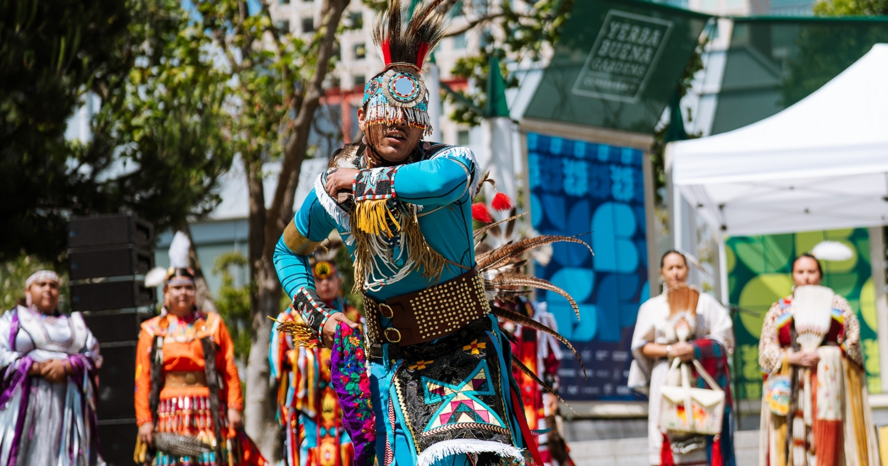 A Native dancer dressed in colorful traditional regalia, demonstrating a dance outdoors, a line of other dancers and the YBG Festival stage behing them.