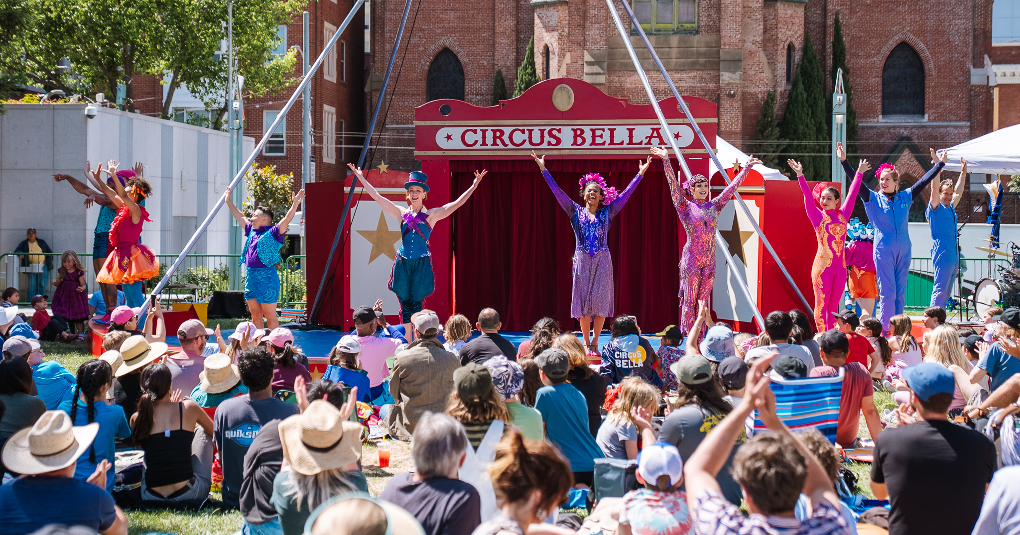Performers of Circus Bella standing across an outdoor stage, wearing colorful costumes, smiling and taking a bow in front of a large audience seated on the grass