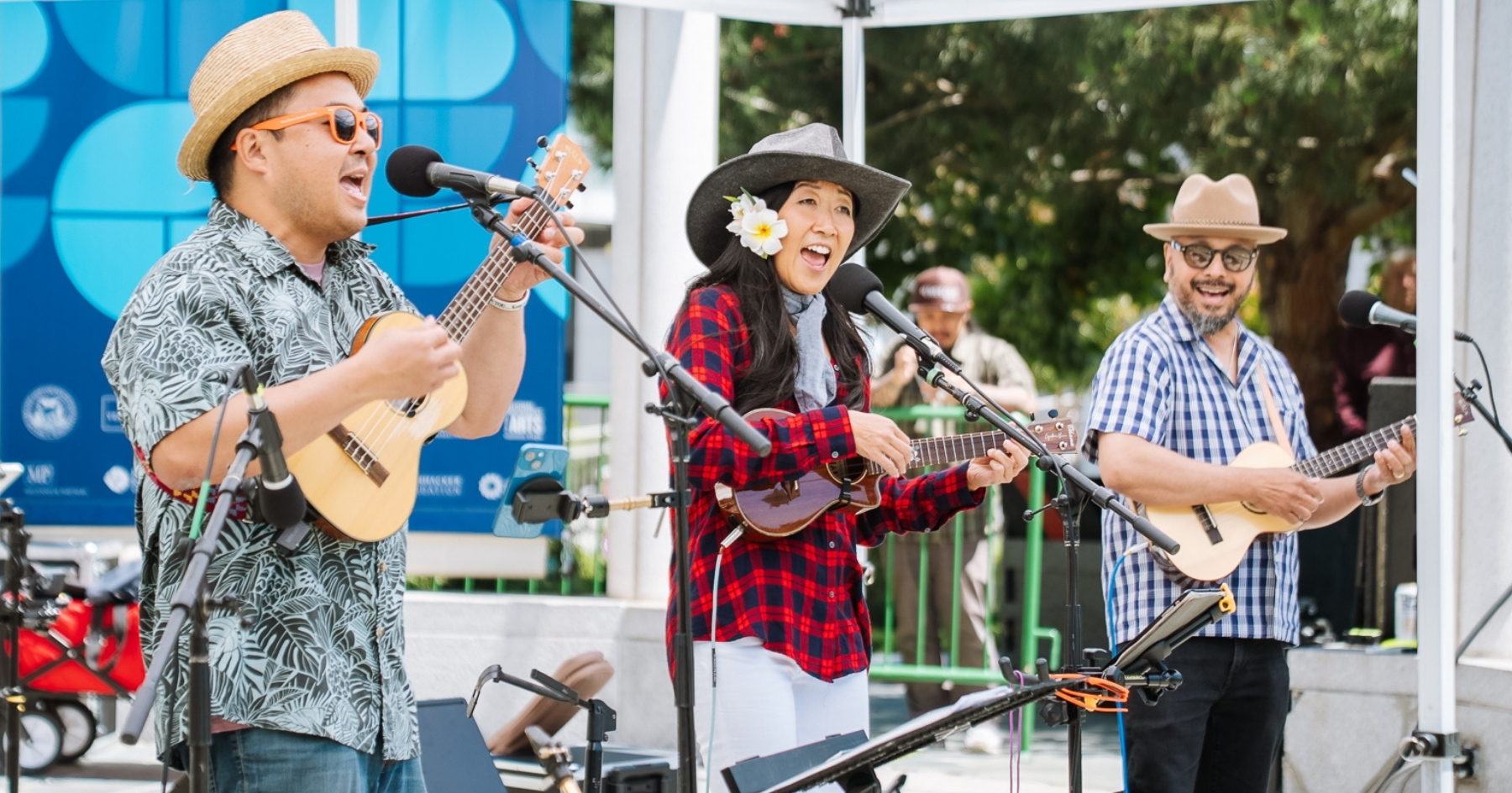 Ukulenny, Cynthia Lin and Steven Espaniola all wearing hats, playing ukuleles and singing into microphones on an outdoor stage.