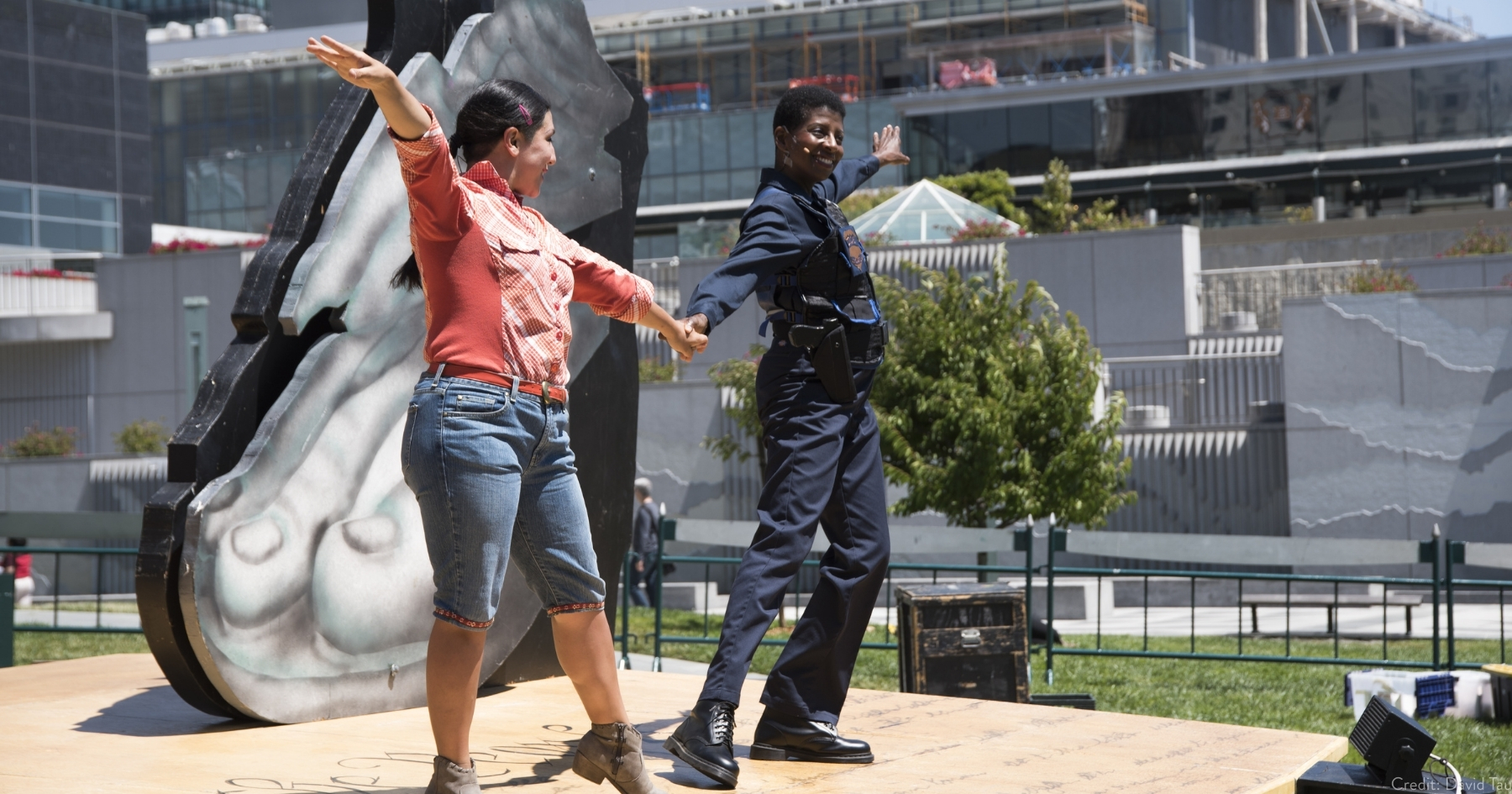 Two actors smiling and holding hands, arms stretched out on a sunny outdoor stage.