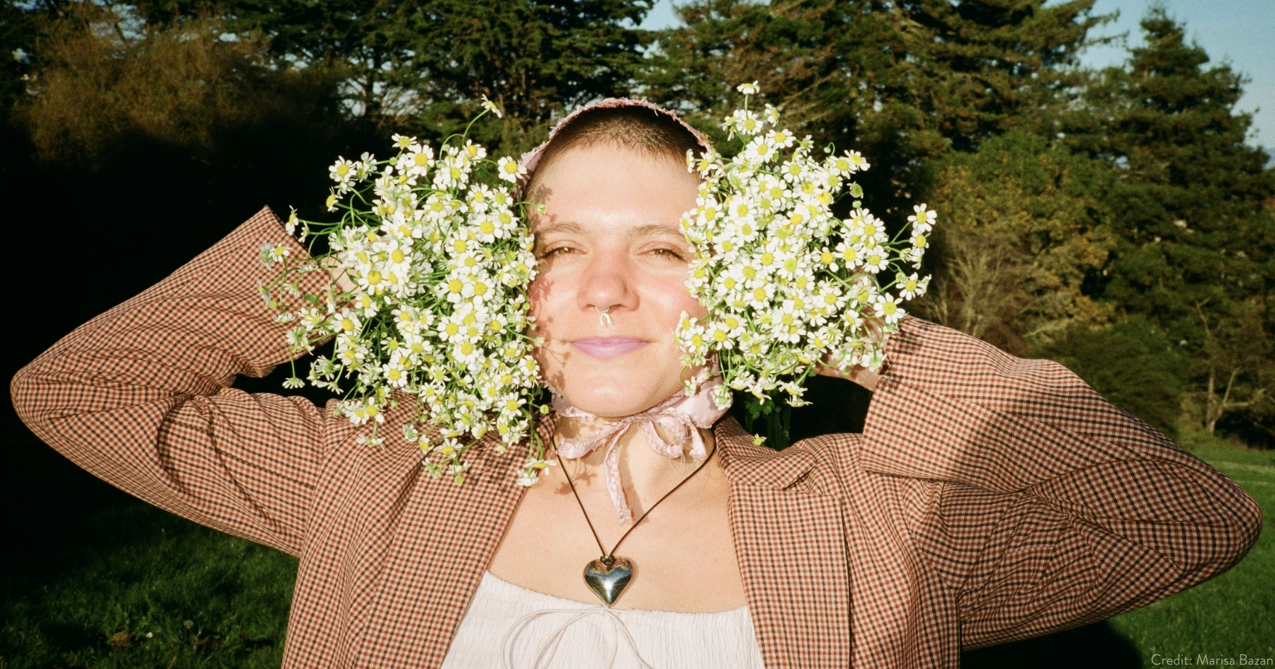 Mae in a white top under a tan blazer, holding up a bunch of white flowers on each side of her head