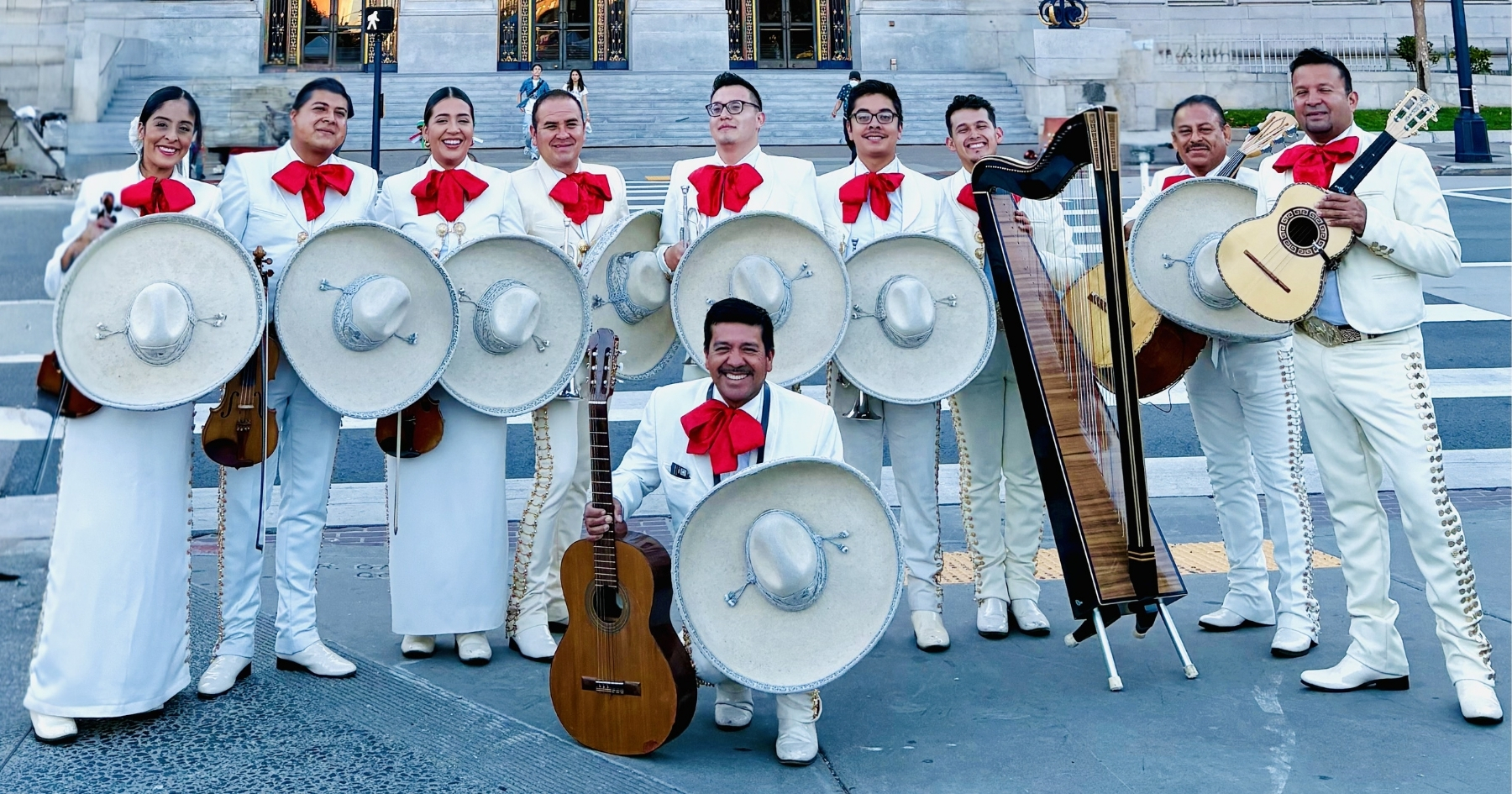 A group of 10 musicians wearing white Mariachi attire with white scarves, posing with various musical instruments