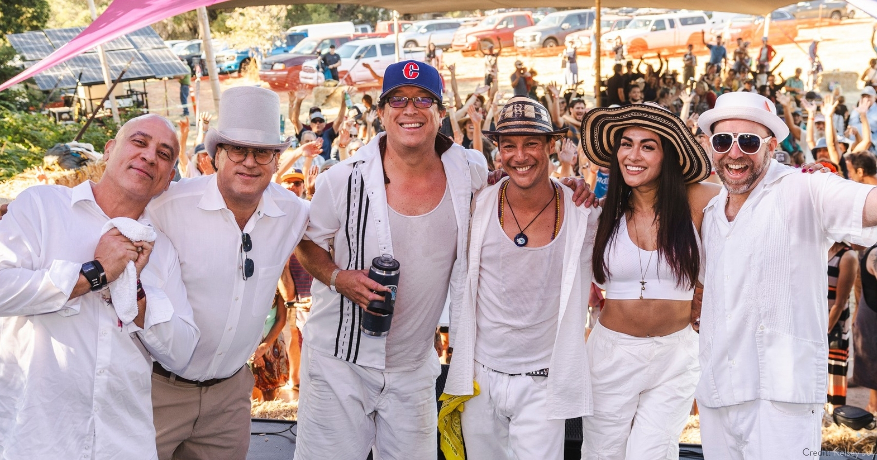 a group of 6 musicians wearing white, smiling shoulder to shoulder with a crowd in the background