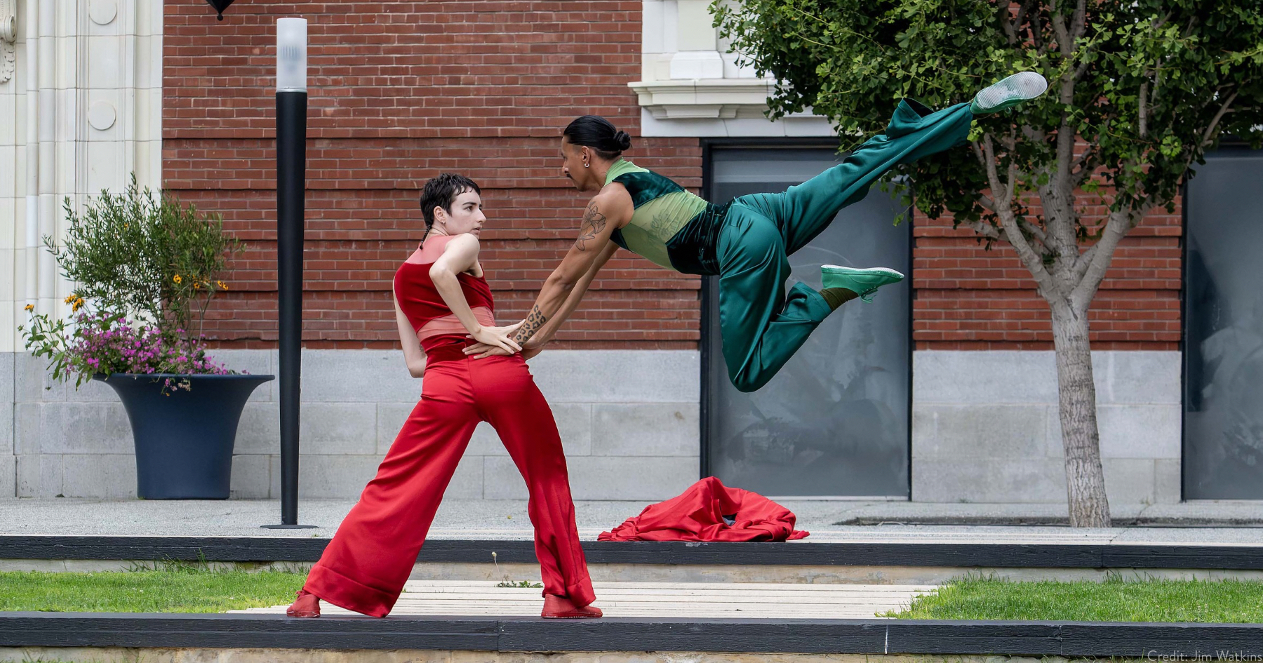 Two dancers in satin red and green costume, dancing outdoors on a brick path on a lawn, a brick building behind them