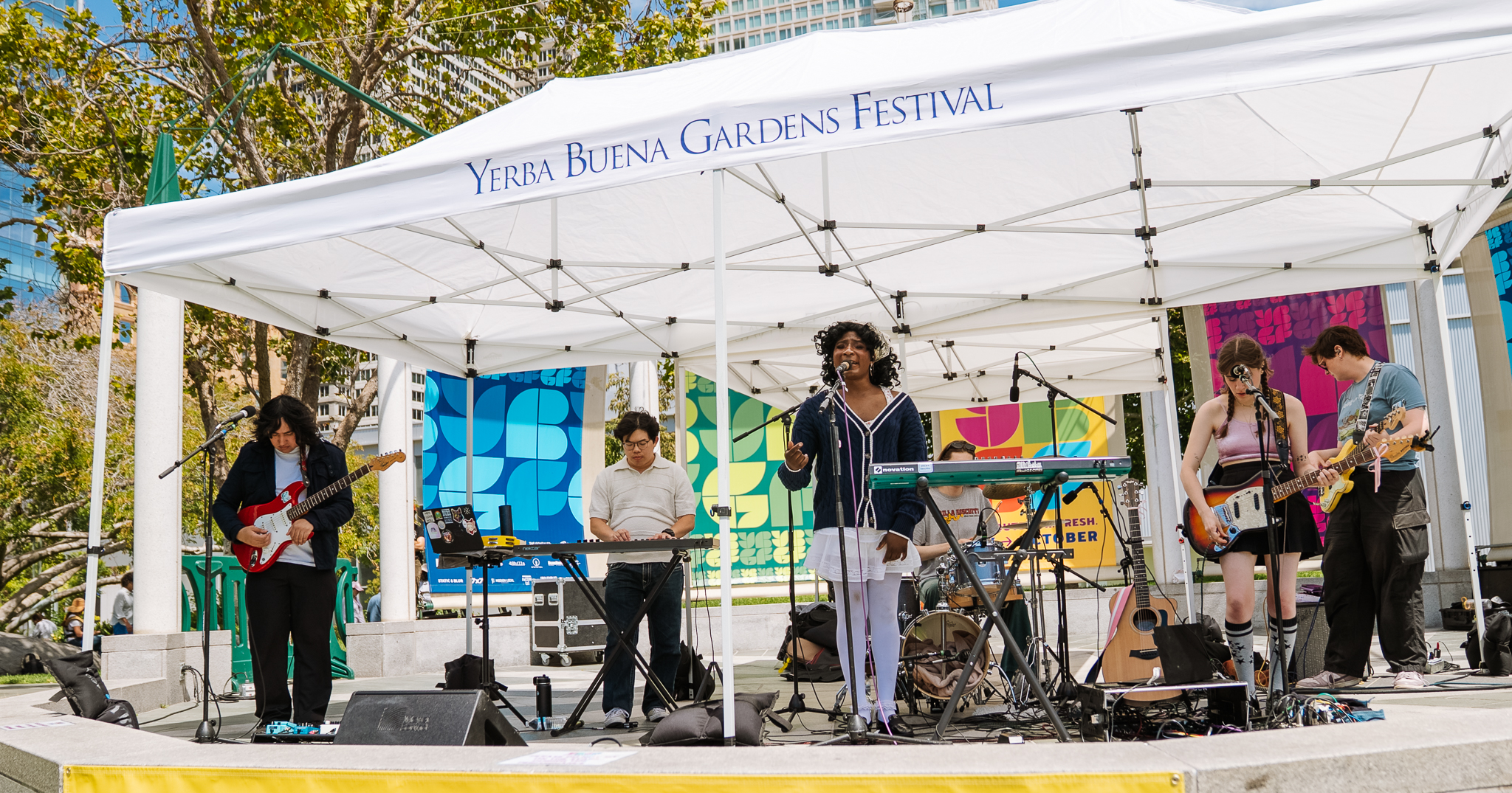 Musical band Mare performing on an outdoor cement stage, covered by a white tent that says "Yerba Buena Gardens Festival"