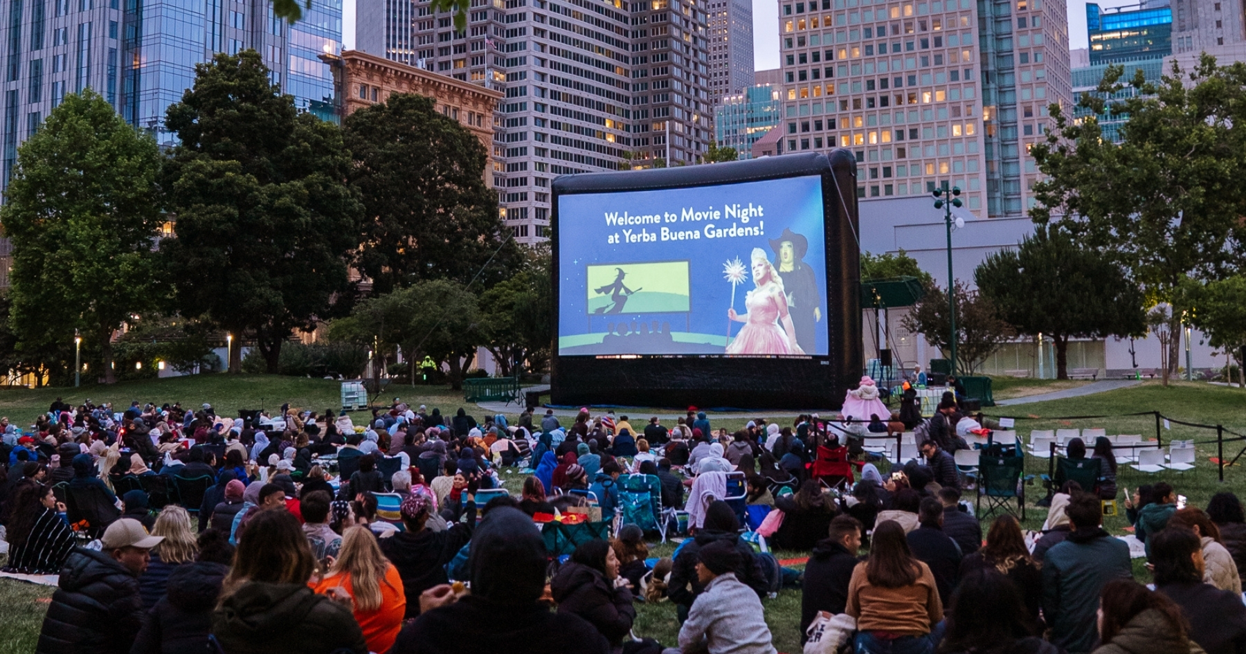 A large audience seated on an outdoor lawn in the dark evening light, watching a movie projected on a large screen, a brightly lit cityscape behind the screen and a line of trees.