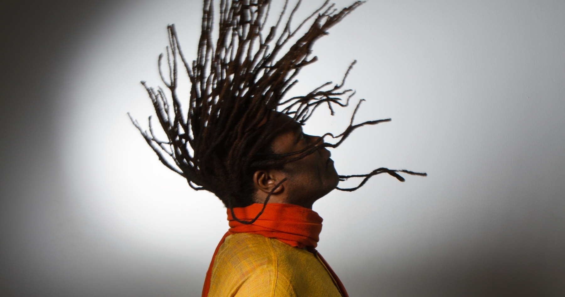 Profile view of Ricardo Lemvo from the shoulders up, earing a yellow top and orange scarf, his head tilted slight up and his long dreadlock hair swung up in the air, a shadow cast on the wall behind him.