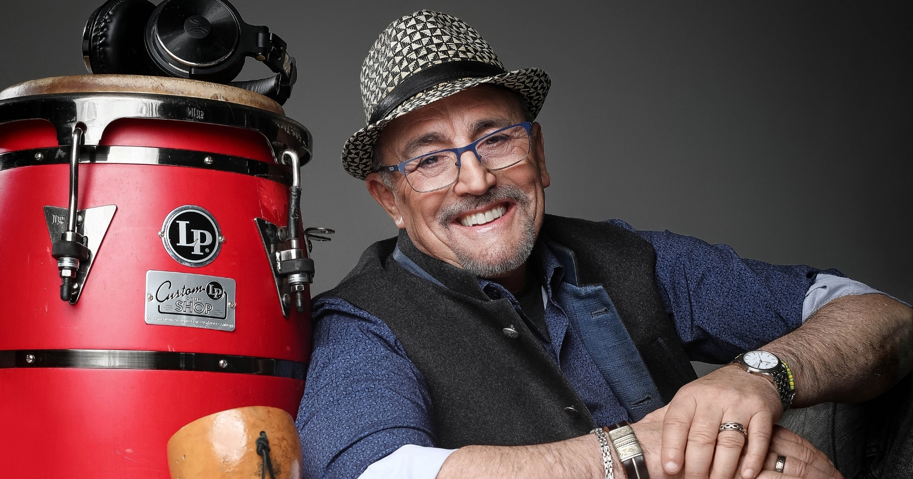 Edgardo Cambón wearing a grey straw fedora hat, glasses, dark blue shirt and grey vest, smiling, leaning to the side agains a bright red conga drum with black headphones on top of it.
