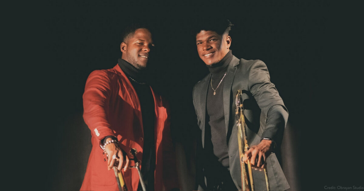 090326_LosCaldarones_1800x945_web2 Obrayan Calderon smiling, wearing a red suit standing next to Dayron Calderon wearing a black suit, each holding out a trombone in one hand, smiling in front of a black background.