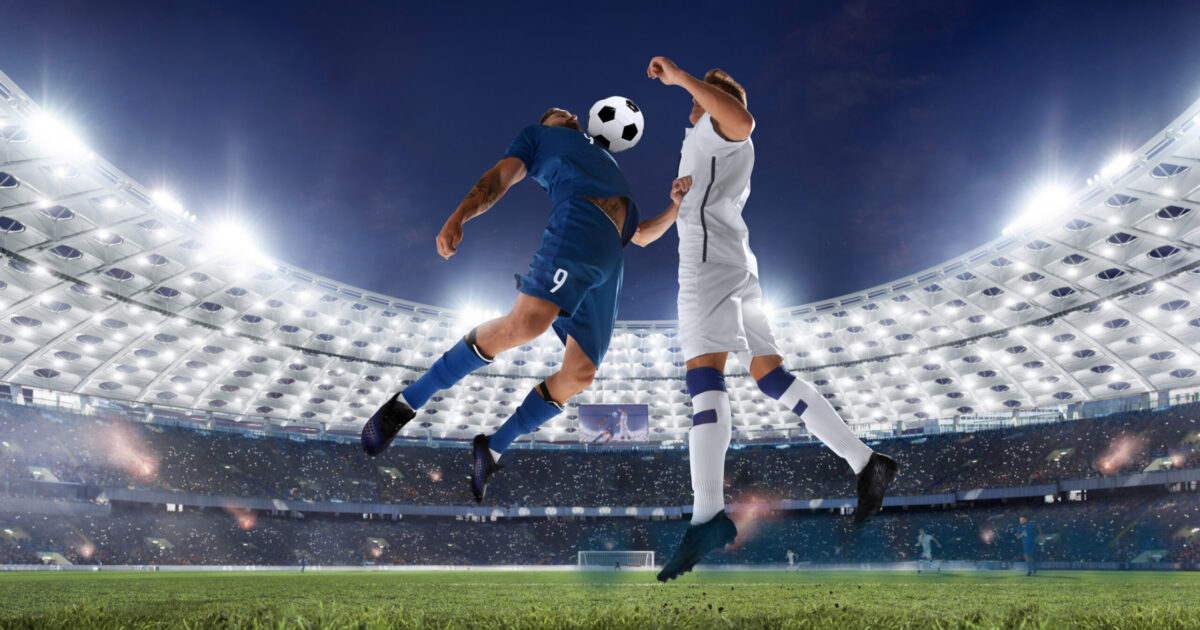 071926_SoccerWatchParty_1800x945-partners-notext Photo of two soccer/football players jumping in the air with ball between their chests, in large, brightly lit soccer/football stadium