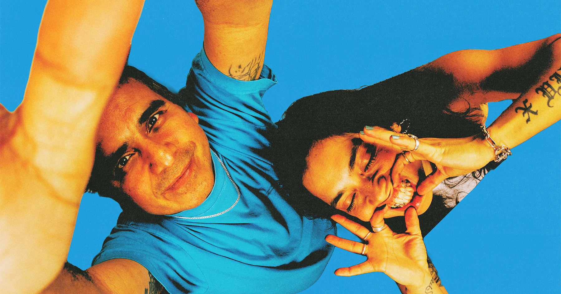 Helado Negro and Reyna Tropical facing up, arms raised up and partially obscuring their faces, in front of a bright blue backdrop